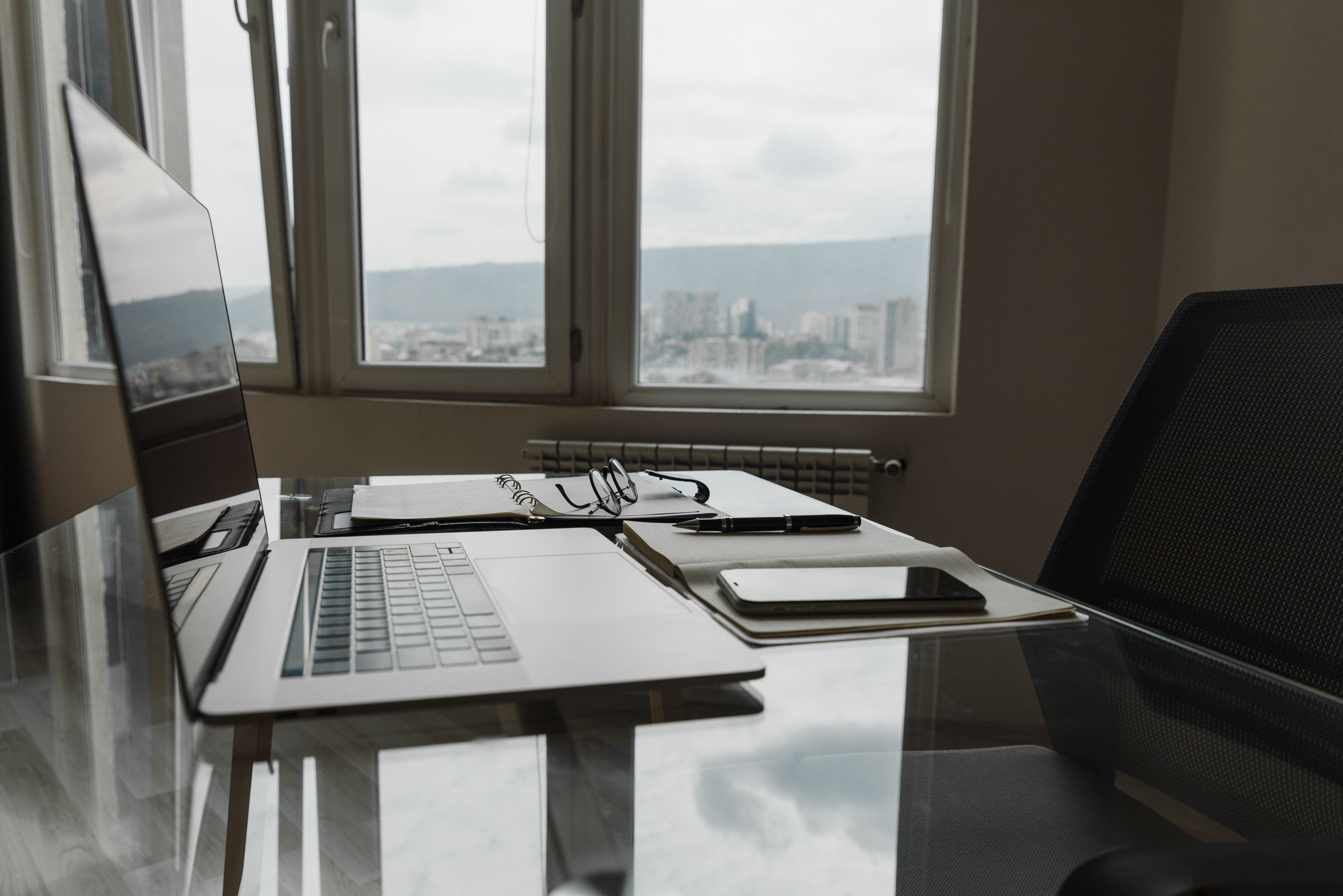 Photograph of a tidy law office workspace with a laptop, notebook and eyeglasses on a glass desk beside a window