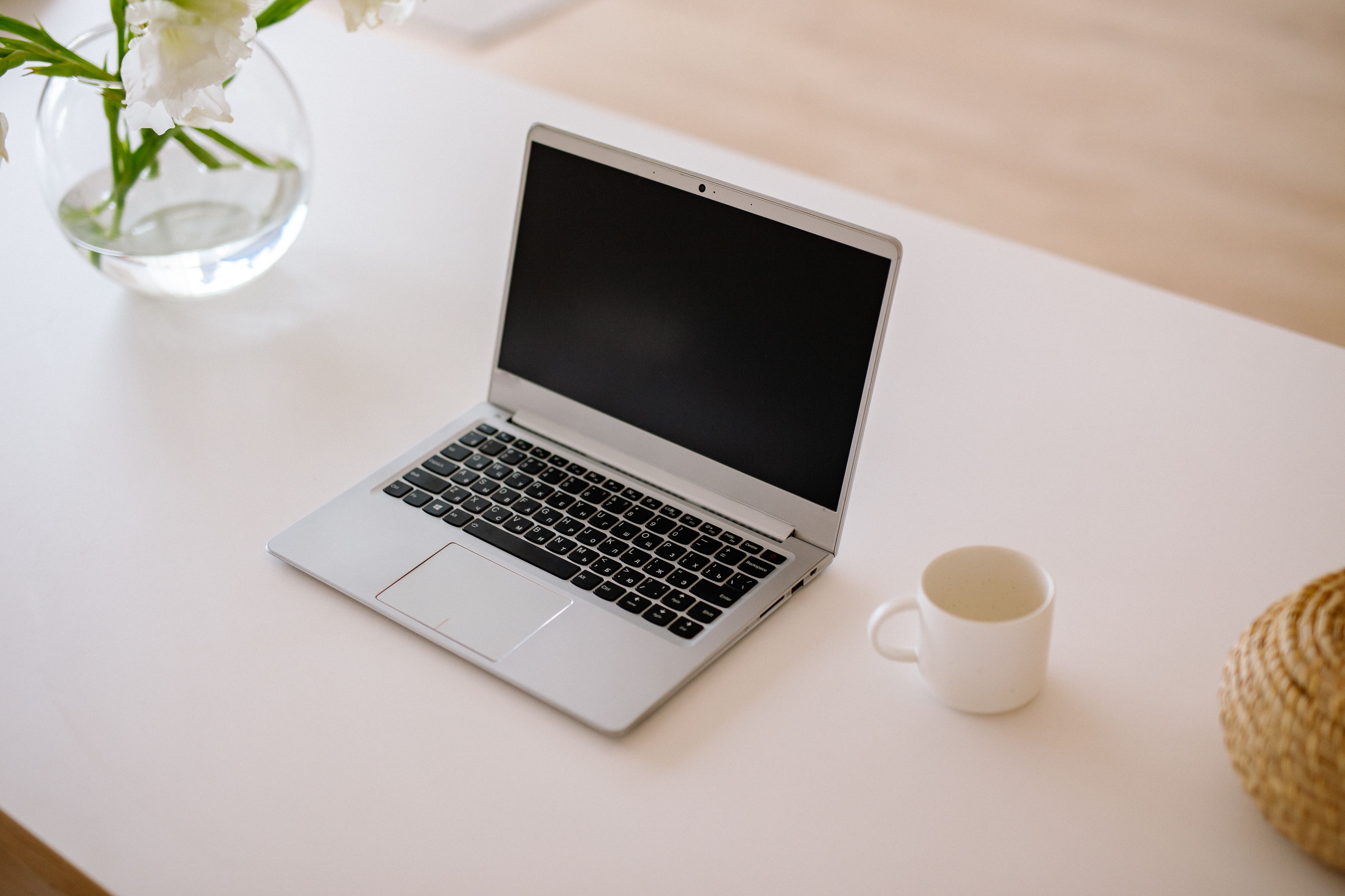 A clean, minimal white desk with an open laptop, a coffee mug and a vase of white flowers