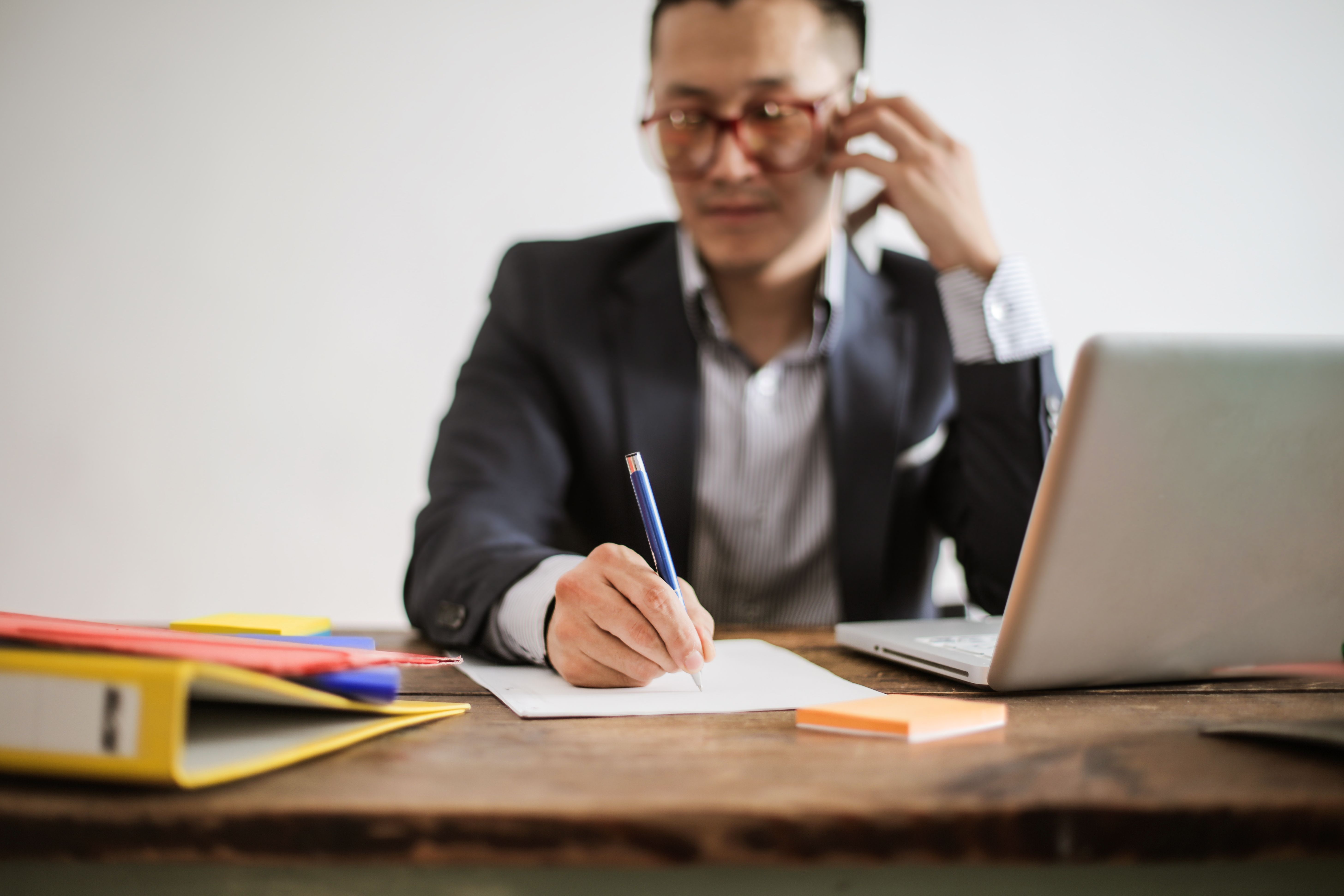 A professional in a suit at an office desk taking notes during a phone call